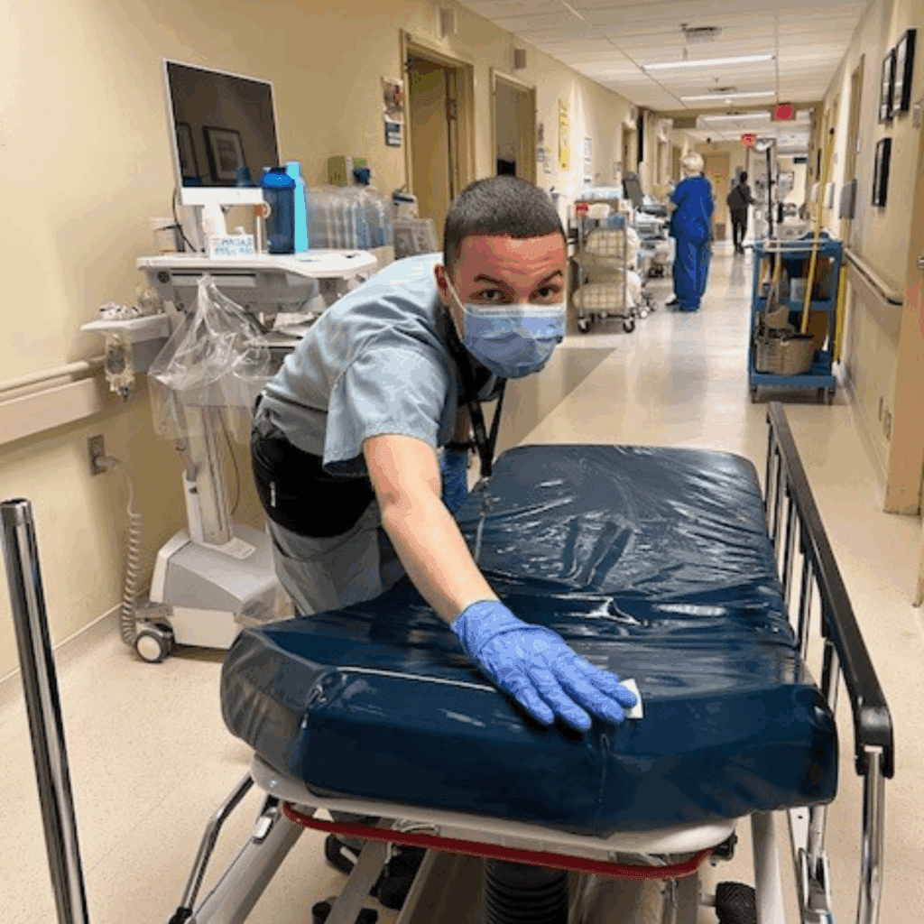 Danial in scrubs in a hospital hallway wearing a mask, gloves, and sanitizing the bed.
