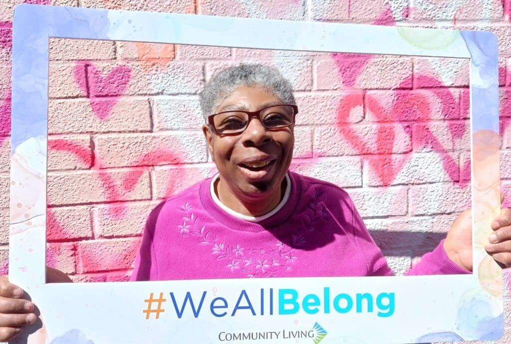 Smiling woman in a pink sweater holds a “#WeAllBelong” frame in front of a heart-painted brick wall.