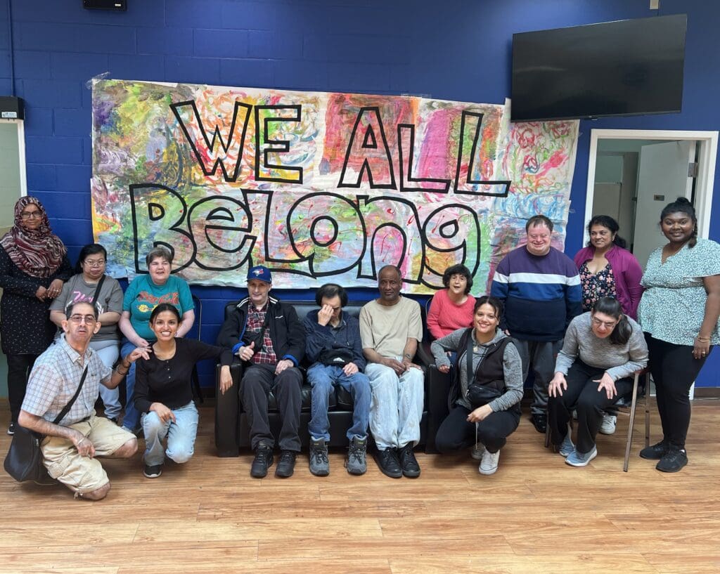 Group of people posing in front of a colorful ‘WeAllBelong’ sign indoors.