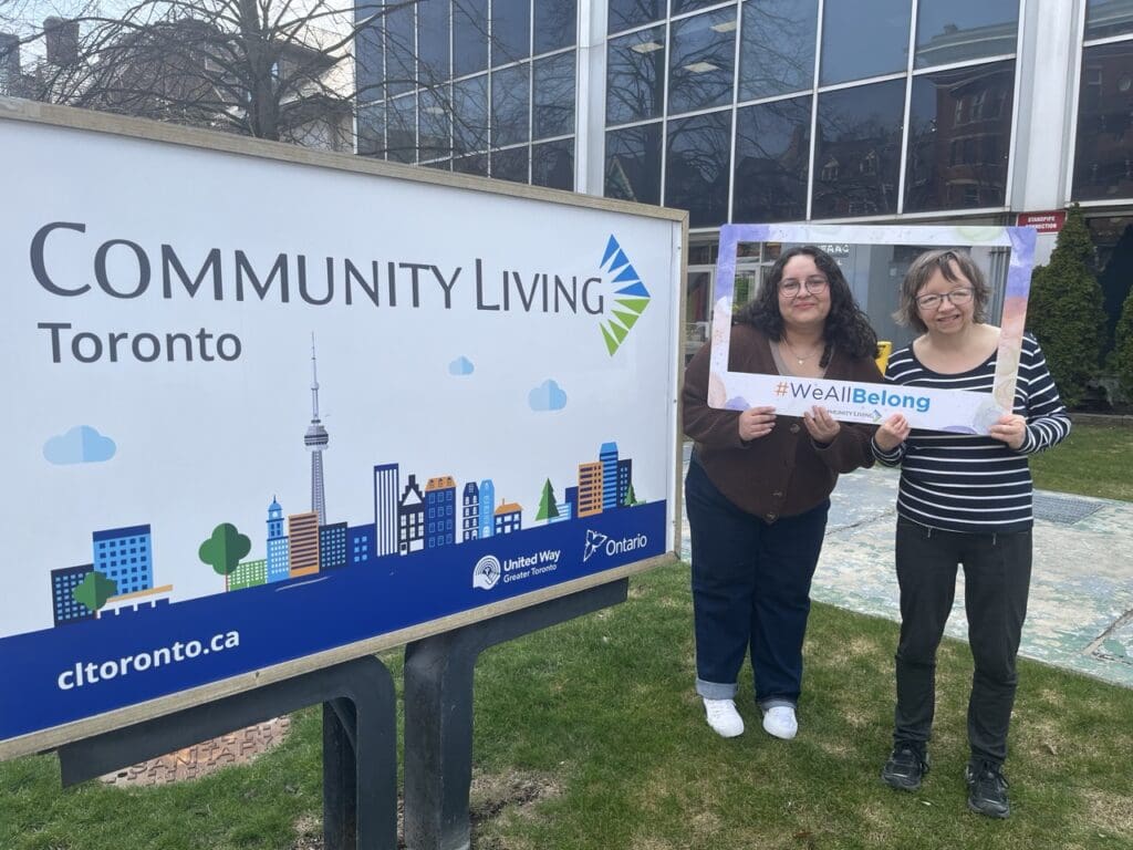Two people holding a ‘#WeAllBelong’ frame beside a Community Living Toronto sign.