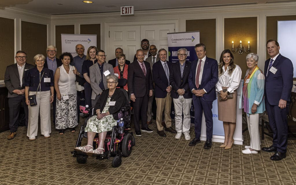 A large group of patrons, board members, and staff stand together indoors for a group photo in front of Community Living Toronto banners. One person is seated in a wheelchair at the front, while others stand behind. Everyone is dressed in formal or semi-formal clothing.