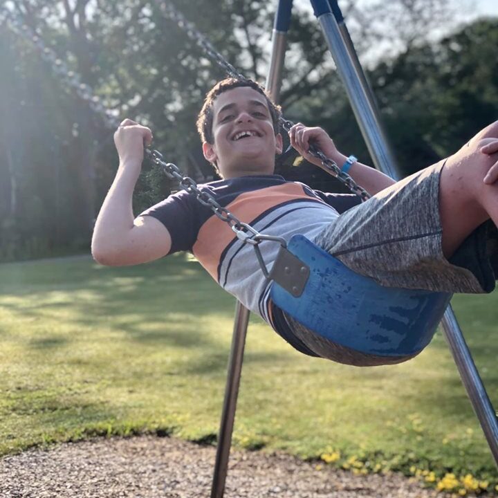 A 12 year old boy on a swing in the summer time. Sunray's visable in the background with tree branches.