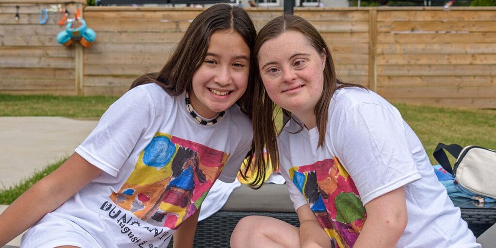 2 smiling young women siting leaning towards each other wearing the same white T-shirt with artwork of a colourful farm, Fence in background